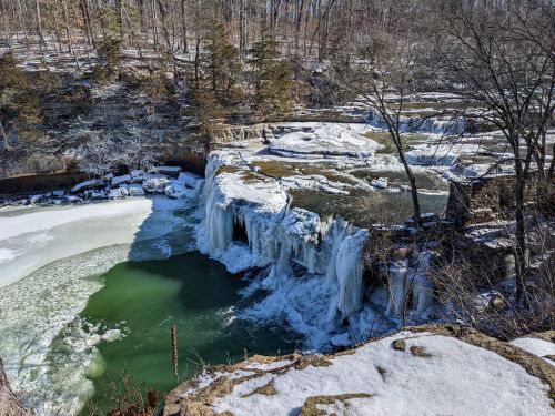 Frozen waterfall cascading into a green pool, surrounded by snow-covered rocks and trees in a winter landscape.