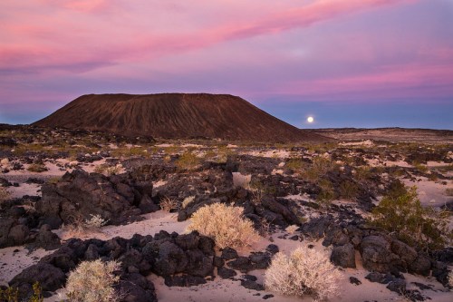 A volcanic cone rises against a colorful sunset sky, with rocky terrain and sparse vegetation in the foreground.