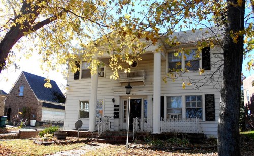 A two-story white house with a porch, surrounded by autumn leaves and trees, in a residential neighborhood.