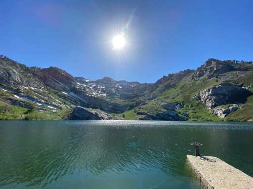 A serene mountain lake surrounded by green hills and rocky cliffs, with a bright sun shining overhead.