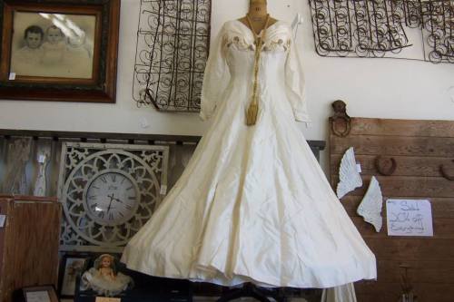 A vintage white dress displayed in a rustic shop, surrounded by decorative items and framed pictures.