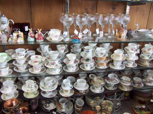 A display of various china teacups, saucers, and glassware on shelves, showcasing intricate floral designs.