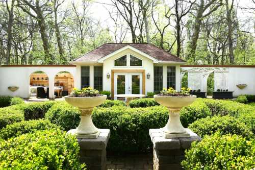 A charming garden with neatly trimmed hedges, two stone planters, and a cozy house surrounded by trees.