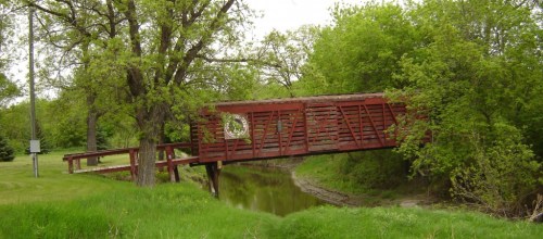 A red wooden bridge spans a calm creek, surrounded by lush green trees and grass.