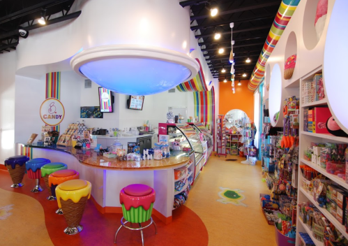 Brightly colored candy shop interior with a curved counter, ice cream-themed stools, and shelves filled with sweets.