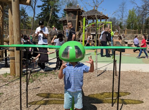 A child in a green helmet plays at a busy playground, surrounded by people enjoying the sunny day.