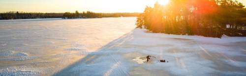 A serene winter landscape with a frozen lake, sunlight glistening, and a person fishing on the ice.