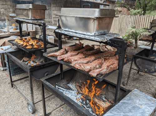 Grills with various meats cooking over open flames in an outdoor setting.