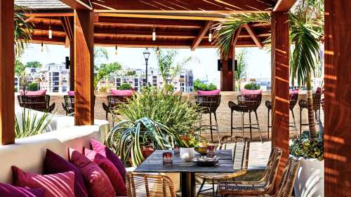 A cozy outdoor dining area with wooden beams, colorful cushions, and lush plants, overlooking a vibrant cityscape.