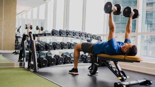 A person lifts dumbbells while lying on a bench in a modern gym with a rack of weights in the background.