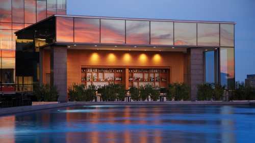 Rooftop bar with a reflective glass facade, surrounded by water and plants, under a colorful sunset sky.