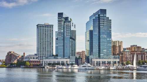 Modern skyscrapers reflect in the water, with boats docked in the foreground and a clear sky above.