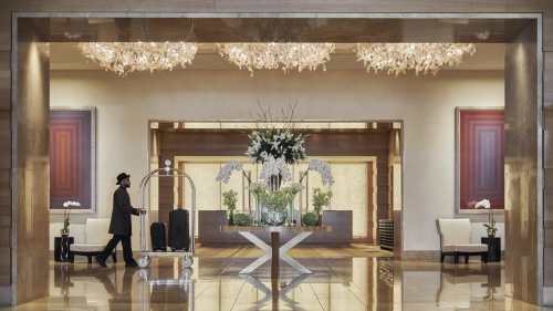 A luxurious hotel lobby with elegant chandeliers, floral arrangements, and a guest with luggage walking by.