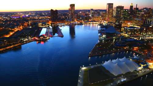 Aerial view of a city skyline at dusk, reflecting lights on a calm river with boats and a large tent on the shore.