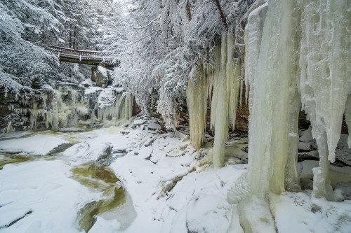A winter scene featuring frozen waterfalls and icicles, surrounded by snow-covered trees and a wooden bridge above.