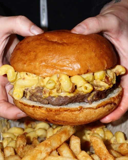A person holds a large burger filled with macaroni and cheese, accompanied by a side of fries.