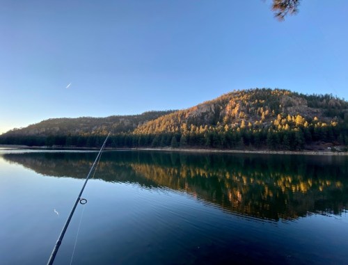 A tranquil lake scene with a fishing rod in the foreground and a forested hillside reflecting on the water.