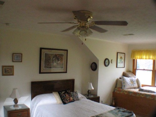 A cozy bedroom featuring a bed, ceiling fan, artwork on the walls, and a window seat with cushions.