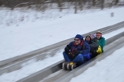 A group of four people joyfully sledding down a snowy track, surrounded by winter scenery.