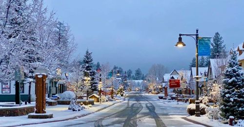 A snowy street lined with trees and quaint buildings, illuminated by streetlights under a cloudy sky.