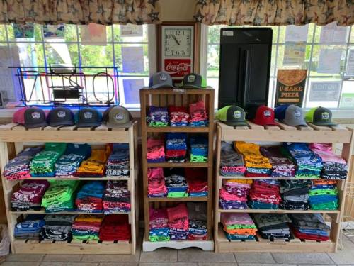 Display of colorful t-shirts and hats arranged on wooden shelves in a shop, with a clock and signs in the background.