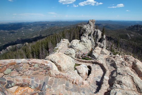 A rocky landscape with a winding stone path, surrounded by trees and distant mountains under a blue sky with clouds.