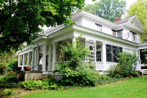 A charming white house with a wraparound porch, surrounded by lush greenery and trees.