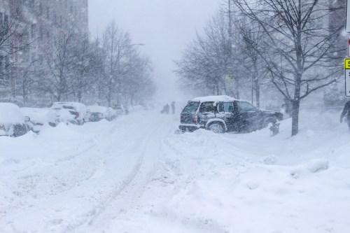 A snowy street with cars buried in snow and people walking in a blizzard, visibility is low due to heavy snowfall.