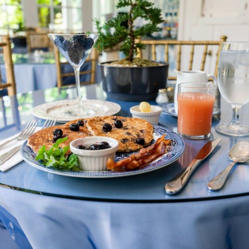 A beautifully set table with a breakfast plate featuring pancakes, blueberries, bacon, and a drink, surrounded by greenery.