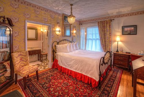 Cozy bedroom with a vintage bed, patterned wallpaper, a rug, and an adjoining bathroom. Warm lighting enhances the ambiance.