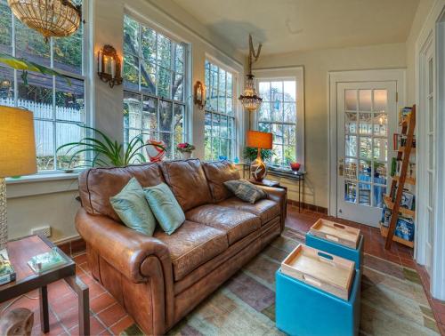 Cozy living room with a brown leather sofa, large windows, plants, and colorful decor, creating a warm atmosphere.