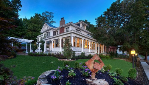 A charming two-story house with a wraparound porch, surrounded by lush greenery and colorful flowers at dusk.