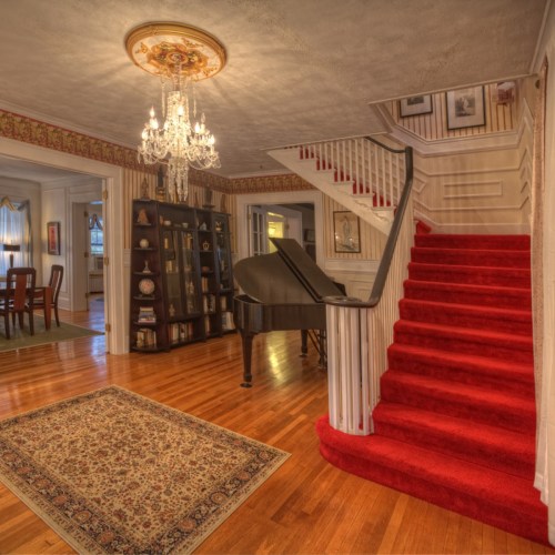 A spacious foyer with a chandelier, red carpeted stairs, a piano, and a patterned rug on hardwood floors.