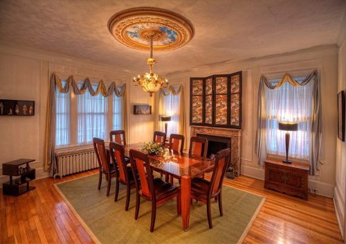 Elegant dining room with a large table, chandelier, and decorative windows, featuring a cozy fireplace and artwork.