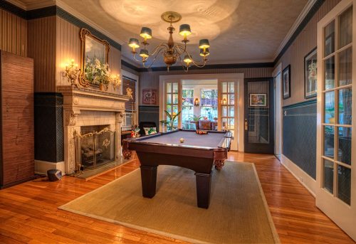 A cozy room featuring a pool table, elegant chandelier, fireplace, and classic decor with wooden flooring.