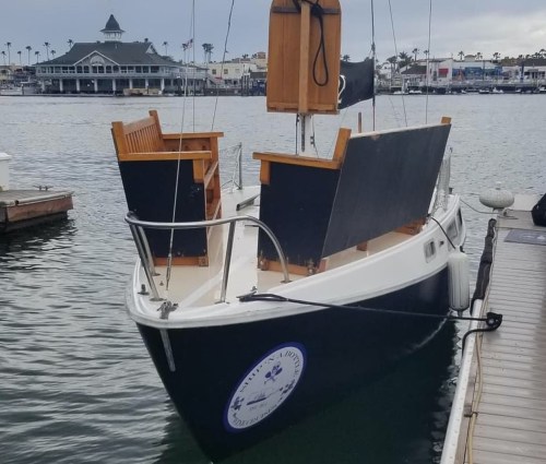 A small boat with wooden seating and a black hull docked in a marina with buildings in the background.