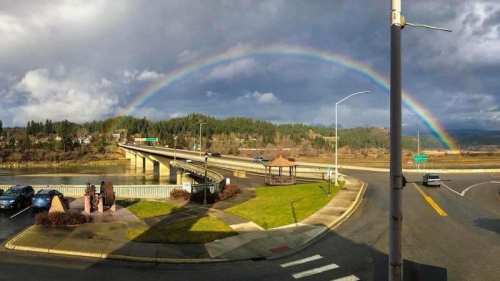 A vibrant rainbow arcs over a highway and green hills, with a gazebo and parked cars in the foreground.