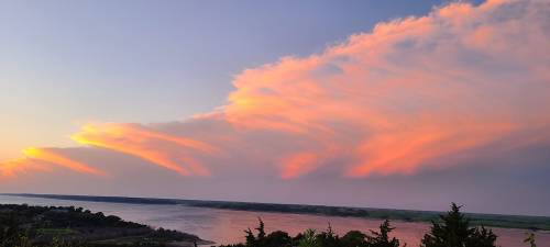 A serene sunset over a river, with soft pink and orange clouds reflecting in the sky. Green trees line the riverbank.