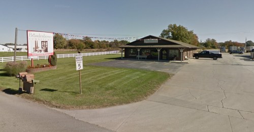 A small building with a sign, surrounded by grass and a few vehicles parked outside, in a rural setting.
