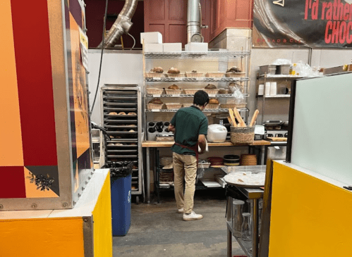 A person in an apron stands at a kitchen counter, surrounded by baking tools and shelves of baked goods.