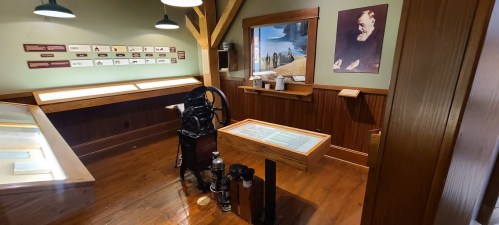 Interior of a museum room featuring a vintage printing press, display tables, and historical images on the walls.