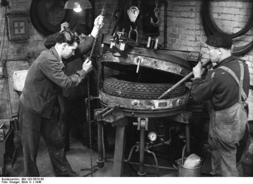 Two men working on a tire machine in a workshop, one operating the machine while the other assists.