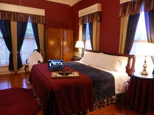 Cozy bedroom with a large bed, wooden furniture, a laptop on a tray, and warm red walls. Soft lighting from lamps.