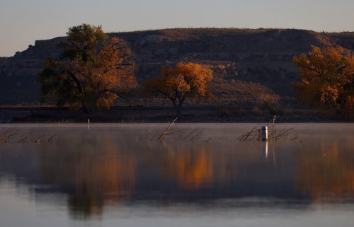 Serene landscape with autumn trees reflecting in calm water, surrounded by mist and distant hills.