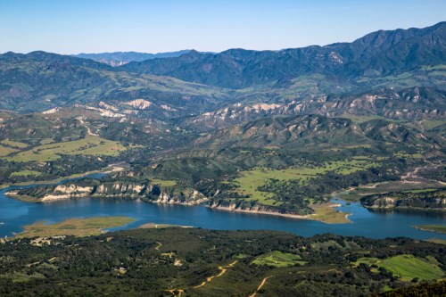 Aerial view of a serene lake surrounded by green hills and mountains under a clear blue sky.