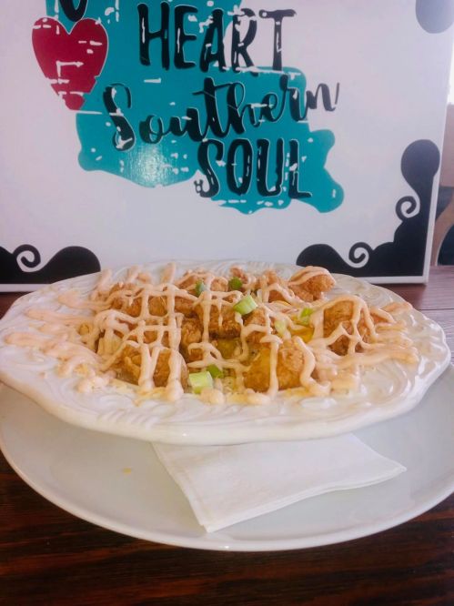 A plate of fried food drizzled with sauce, garnished with green onions, in front of a colorful sign that reads "Heart Southern Soul."