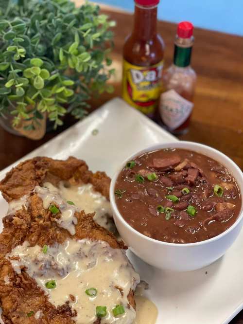 A plate of fried chicken with gravy, served alongside a bowl of beans topped with green onions and hot sauce bottles in the background.