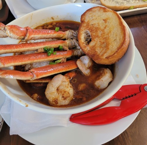 A bowl of seafood stew with shrimp and crab legs, accompanied by a slice of toasted bread and a red seafood cracker.