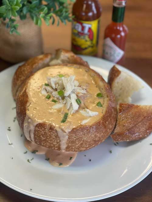 A bread bowl filled with creamy soup, topped with shredded chicken and green onions, served with hot sauce bottles.