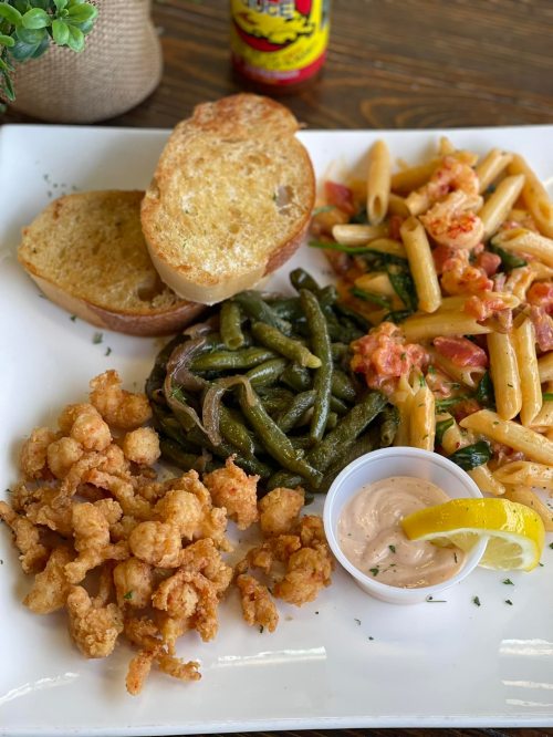 A plate featuring pasta, green beans, fried shrimp, toasted bread, and a dipping sauce with a lemon wedge.